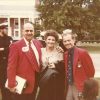 Two white men in red jackets with white woman smiling together with sailors in uniform and brick building behind them