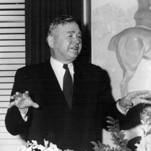 white man in suit and tie gestures while speaking  in front of painting and window obscured by curtains, blinds