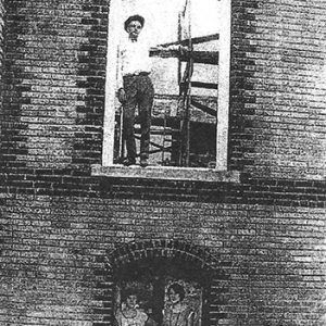 Brick church under construction with white man standing in upper story window frame and two white women looking out the bottom story window frame