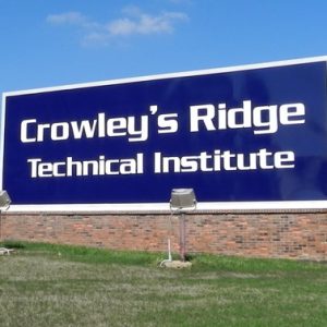 Large blue sign with white text and lights on grass with Brick building with parking lot behind it