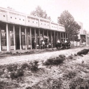 Group of white men standing and two white men on horseback at brick storefront with covered walkway on dirt road