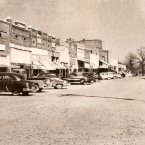 Multistory brick storefronts and parked cars on dirt road