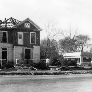 Two-story house with roof partially destroyed sitting on street with other debris visible and other structures in the distance