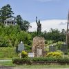 Statue of Native American man on stone pedestal in flower bed with historical markers surrounding it and gazebo on hill in the background