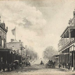 Horse drawn carriages on dirt road with multistory buildings on both sides with covered entrances