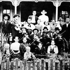 Group of white musicians with their instruments accompanied by white women and children inside fence with house behind them