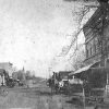 Horse drawn carriages on town street with multistory building with tower in the background