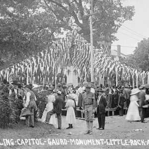 Crowd of people gathered around monument with statue and flags hanging above them