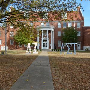 Multistory brick building with round covered porch with columns and Greek letters in front yard with trees