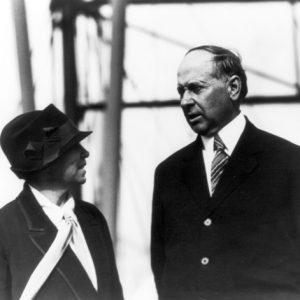 White woman in ribbon hat suit and tie speaking to white man in suit outdoors