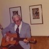 Old white man in suit and glasses playing an acoustic guitar on couch