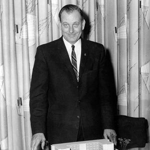 White man smiling in suit and tie with building model on table