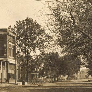 View looking down street towards multistory building with storefronts on one side and trees running along the other side