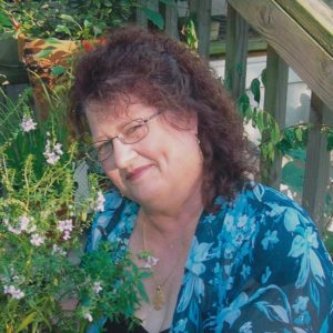 White woman with long hair and glasses in blue floral top surrounded by plants