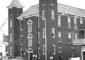 Three-story brick building with twin bell towers and street with parked cars