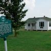 Single-story house with white paneling and historical marker sign on grass with tree