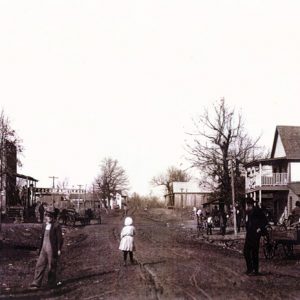 White men in overalls and little girl in white dress standing in dirt road with stores and trees on both sides