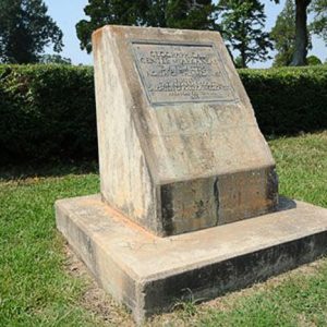 Concrete monument with base and plaque and hedge behind it