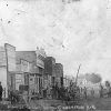 White men women and children on horse drawn wagons on dirt road with brick storefronts and church in the background