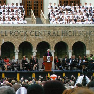 white man in suit and red tie on dais with other individuals, white and African American, in front of brick building labeled "Little Rock Central High School" before a large crowd