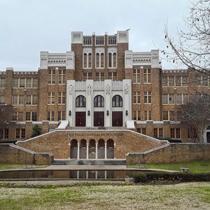 Multistory brick school building with twin staircases leading to front with pool and trees in foreground