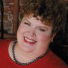 White woman with curly hair smiling in red top with bead necklace