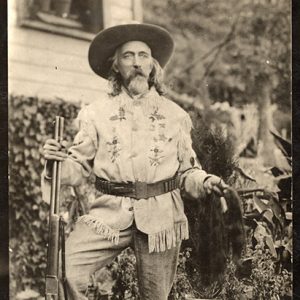 Old white man with long hair and beard in western clothing with hat and gun