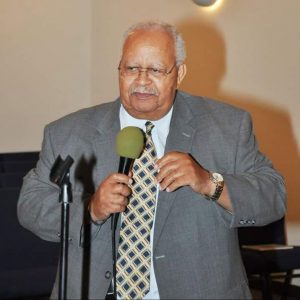 Old African-American man in suit and tie speaking into microphone in church sanctuary