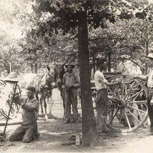 Group of men outdoors next to wooden structure working on wagon wheels and frames and one man with two donkeys behind them