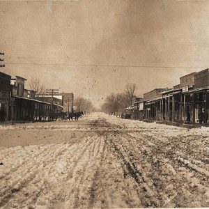 Dirt town street with storefronts on both sides and horses in the middle distance