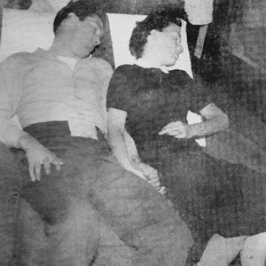 White man in shirt and pants and woman in a dark dress holding hands and sleeping in train car