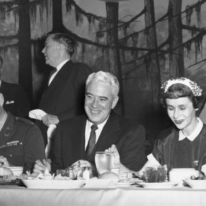 White men in suits and military uniform and woman eating a banquet with fake forest scene behind them