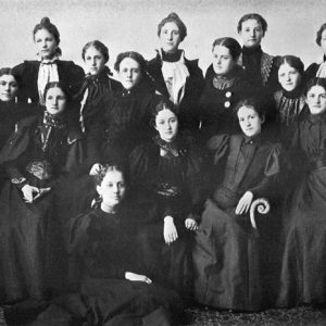 Group of more than a dozen white women posing together in black dresses and pulled-back hair