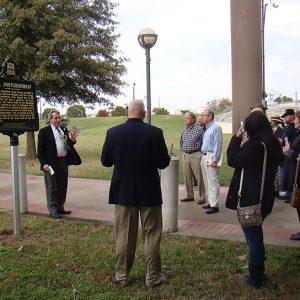 White man in suit and tie standing next to sign speaking to group