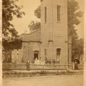 White men and women gathered outside brick church building with tall central tower and fence on dirt road