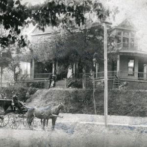 Funeral wagon and horses on street with people watching from their elevated multistory house across the street