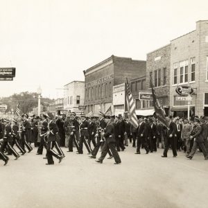 Downtown parade with marching band soldiers spectators brick buildings "rexall drugs westinghouse coca cola."