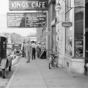 White residents walking on sidewalk by storefronts under signs past parked cars