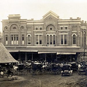 multi story "John C Hill 1883" brick building dirt road gazebo cars horse carriages audience