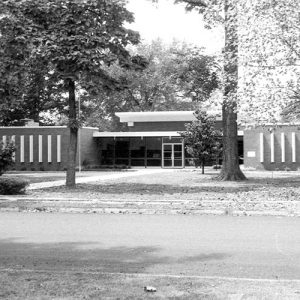 Single-story building with large glass windows around the entrance surrounded by tall trees