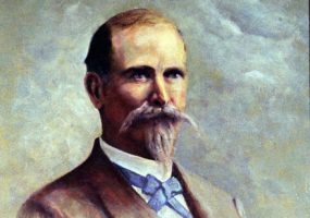 White man with handlebar mustache and beard in suit and blue bow tie