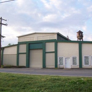 Green and white warehouse on street with power lines and water tower behind it