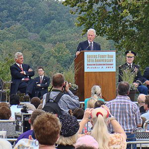 White man with white hair speaking into a microphone at lectern to a crowd with white men in suits and military officer on stage