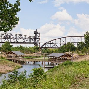 Steel truss bridge over river behind wooden pavilions connected by wooden walking bridge