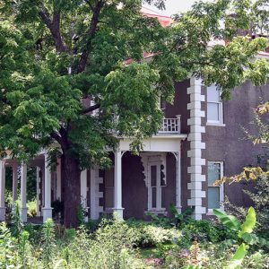 Two-story house with porch and decorative brick corners and tower behind a tree with gardens