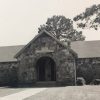 Single-story building with stone walls covered porch and arched entrance on grass with walking path