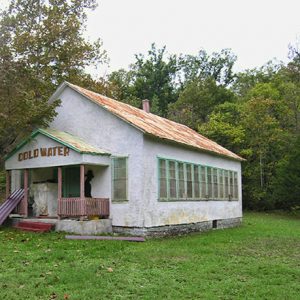 rundown white building with green trim with "Cold Water" painted above the entrance and a black silhouette on the porch