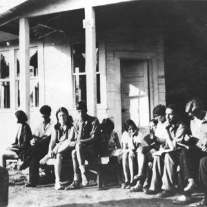 White man sitting in front of white students on porch of wooden building