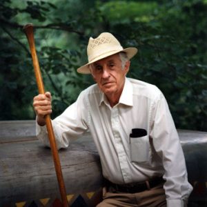 Old white man in hat holding a paddle posing with canoe and trees in the background