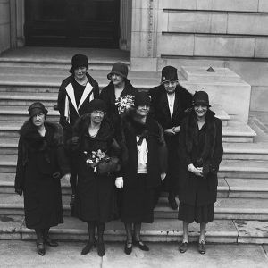 Group of seven white women standing on marble stairs in dresses coats and hats